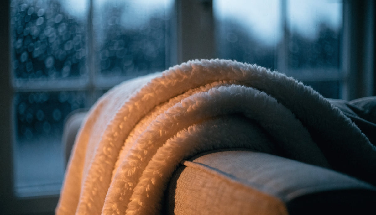 A soft cream fleece blanket folded over a sofa arm in warm lamplight on a winter evening